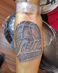 Фото татуировки баскетбольного мяча с надписью "True to the Game" на предплечье. Черно-белый рисунок татуировки в стиле графика.