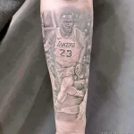 Фото тату с изображением Леброна Джеймса в форме Los Angeles Lakers на предплечье, выполненное в реалистичном стиле с использованием черно-серых оттенков.
