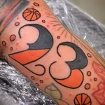 Фото яркой татуировки на руке с изображением номера 23 в оранжевом цвете, окруженного баскетбольными мячами и брызгами краски.