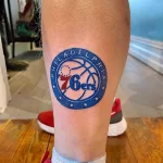 Фото тату красочного логотипа баскетбольной команды Philadelphia 76ers с баскетбольным мячом и звездами, выполненное на икре.