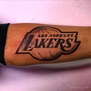 Фото тату на руке с изображением логотипа баскетбольной команды Los Angeles Lakers, выполненное в черном цвете.