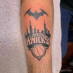 Фото тату на руке, сочетающее логотип баскетбольной команды New York Knicks с силуэтом Бэтмена и изображением небоскребов Нью-Йорка. Черно-белая татуировка в стиле графики.