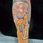 Фото тату на руке с реалистичным портретом Kobe Bryant в желтой форме Lakers с номером 24. Детализированный рисунок татуировки.