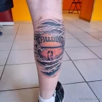 Фото тату на колене с изображением баскетбольного мяча Spalding, с автографом и эффектом трещин. Реалистичный рисунок татуировки.