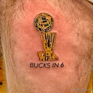 Фото тату на груди с изображением баскетбольного трофея и надписью 'BUCKS IN 6'. Яркий рисунок, символизирующий победу.