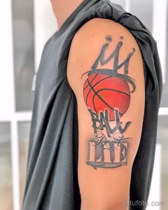 Фото татуировки баскетбольного мяча с короной и надписью 'Ball Don't Lie', выполненной в черно-серых тонах на плече.