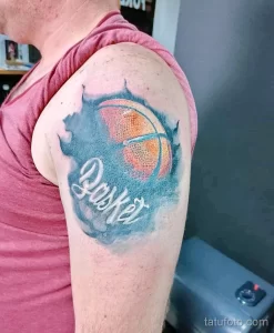 Фото тату баскетбольного мяча с надписью "Basket" на плече. Рисунок выполнен в акварельной технике с использованием синих и оранжевых оттенков.