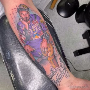 Фото тату на руке, изображающее баскетболиста Коби Брайанта в форме Lakers, держащего мяч. Яркий, цветной рисунок татуировки с надписью "Mamba Mentality".