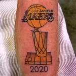 Фото тату с изображением кубка NBA команды Los Angeles Lakers с надписью 2020 года на предплечье. Черно-белый рисунок татуировки.