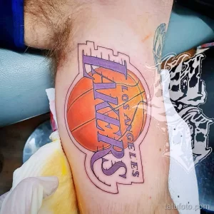 Фото тату на предплечье с изображением логотипа баскетбольной команды Los Angeles Lakers. Рисунок выполнен в ярких цветах с использованием желтого, фиолетового и белого.