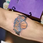 Фото тату на руке с изображением логотипа баскетбольной команды New York Knicks, выполненное в ярких цветах с использованием теней.
