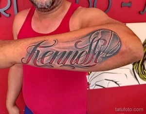 Цветное фото татуировки на руке с надписью "Kenneth" в стильном шрифте с декоративными элементами.