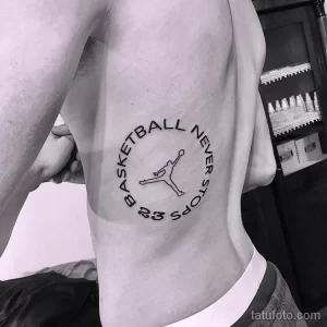 Черно-белое фото татуировки на боку мужчины с надписью "Basketball Never Stops" и силуэтом Майкла Джордана в прыжке.