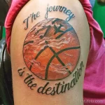 Фото тату баскетбольного мяча с надписью "The journey is the destination" на плече. Яркий рисунок в оранжево-черных тонах.