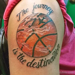 Фото тату баскетбольного мяча с надписью "The journey is the destination" на плече. Яркий рисунок в оранжево-черных тонах.