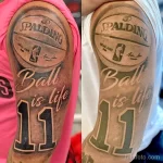 Фото татуировки на плече: логотип Spalding, надпись 'Ball is life' и баскетбольный мяч. Черно-серые тона, реалистичный стиль.