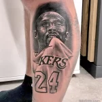 Фото татуировки на ноге с реалистичным портретом Коби Брайанта в форме Lakers с номером 24. Детализированный черно-белый рисунок.