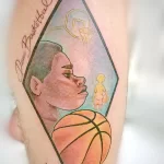 Фото тату баскетболиста в профиль, выполненное в ярких цветах и стиле комиксов на предплечье. Эскиз татуировки включает баскетбольный мяч и кольцо.