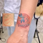 Фото тату небольшого баскетбольного мяча со стрекозой и надписью "twelve" на запястье. Цветная татуировка, яркий дизайн, личное значение.