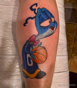 Фото тату на ноге с изображением синего мультяшного персонажа в баскетбольной форме, держащего мяч. Яркие цвета и динамичная композиция.