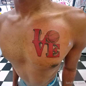 Фото татуировки на груди с надписью "LOVE", в которой буква "O" заменена на баскетбольный мяч. Яркий красный цвет и смелый дизайн.