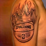 Черно-белое фото тату на плече с изображением баскетбольного мяча Spalding, увенчанного короной и окруженного пламенем. Детализированный рисунок с эффектом теней.