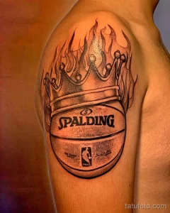Черно-белое фото тату на плече с изображением баскетбольного мяча Spalding, увенчанного короной и окруженного пламенем. Детализированный рисунок с эффектом теней.