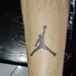 Фото тату с изображением логотипа Jumpman (Майкл Джордан в прыжке). Черно-белый минималистичный рисунок на предплечье.