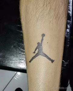 Фото тату с изображением логотипа Jumpman (Майкл Джордан в прыжке). Черно-белый минималистичный рисунок на предплечье.