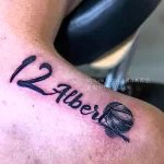 Фото татуировки на плече с изображением баскетбольного мяча и надписью '12 Albert'. Простой и стильный дизайн.