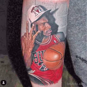Фото тату с изображением Майкла Джордана в форме Chicago Bulls, делающего жест двумя пальцами. Детализированный портрет и баскетбольный мяч.