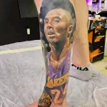 Фото тату с реалистичным портретом Коби Брайанта в форме игрока Lakers на предплечье. Детализированный рисунок татуировки выполнен в черно-серых тонах.