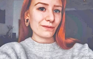 Фото тату: девушка с рыжими волосами, растяжками в ушах и пирсингом в носу. Крупный план лица девушки с улыбкой.