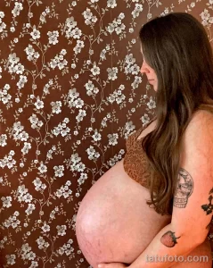 Фото тату на руке беременной женщины на фоне цветочного узора. Рисунок татуировки выполнен в минималистичном стиле, изображает клубнику.