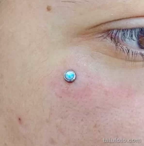 Фото тату: пирсинг под глазом с небольшим голубым камнем украшает кожу девушки.