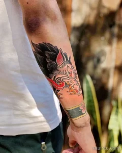 Фото тату: традиционный рисунок головы волка с красными брызгами на предплечье, выполненный в ярких цветах и четких линиях.