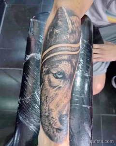 Фото тату волка с голубыми глазами на руке. Реалистичный рисунок в черно-серых тонах с акцентом на выразительные глаза. Татуировка выполнена на предплечье.
