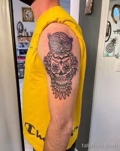Фотография татуировки совы, стилизованной под сахарную черепушку (calavera) с цветочными узорами на плече.