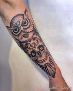 Черно-белое фото тату совы, стилизованной под череп, с элементами чикано на предплечье.