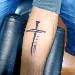 Фото тату креста с надписью 'Joshua 1:9' на предплечье. Черно-белая татуировка, выполненная четкими линиями, символизирует религиозные убеждения и силу духа.