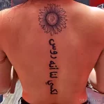 Фото тату подсолнуха с арабской надписью на спине девушки. Черно-белый рисунок, детально проработанные лепестки и изящная каллиграфия.