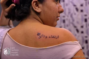 Фото тату с арабской надписью и изображением короны на спине девушки. Черные чернила, каллиграфия, минималистичный стиль.