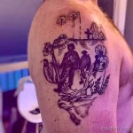 Фото тату на плече с изображением силуэтов двух людей, сидящих среди полевых цветов и креста на горизонте. Черно-белый рисунок татуировки.