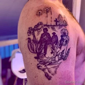 Фото тату на плече с изображением силуэтов двух людей, сидящих среди полевых цветов и креста на горизонте. Черно-белый рисунок татуировки.