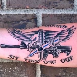 Фото тату на предплечье: автомат, крылья, надпись "Sit This One Out". Военная тематика, выполненная в черно-серых тонах с цветными акцентами.