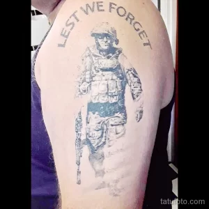 Фото татуировки на плече с изображением солдата и надписью "Lest We Forget". Черно-белый рисунок.