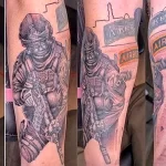 Фото татуировки на ноге, изображающей нескольких солдат в боевой обстановке, военную технику и пейзаж, выполненное в реалистичном стиле с высокой детализацией.