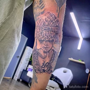 Черно-белое фото тату на предплечье с изображением рэпера 2Pac в характерной шапке и бандане, держащего сигарету. Детализированный рисунок татуировки.