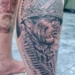 Фото тату на ноге: реалистичное изображение бойца в каске с оружием и гильзами. Детализированный рисунок в черно-серых тонах, символизирующий мужество и героизм.