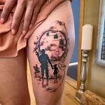 Фото татуировки на бедре с изображением отца и дочери, стоящих под полной луной. Стилизованный рисунок с использованием черных и серых оттенков.
